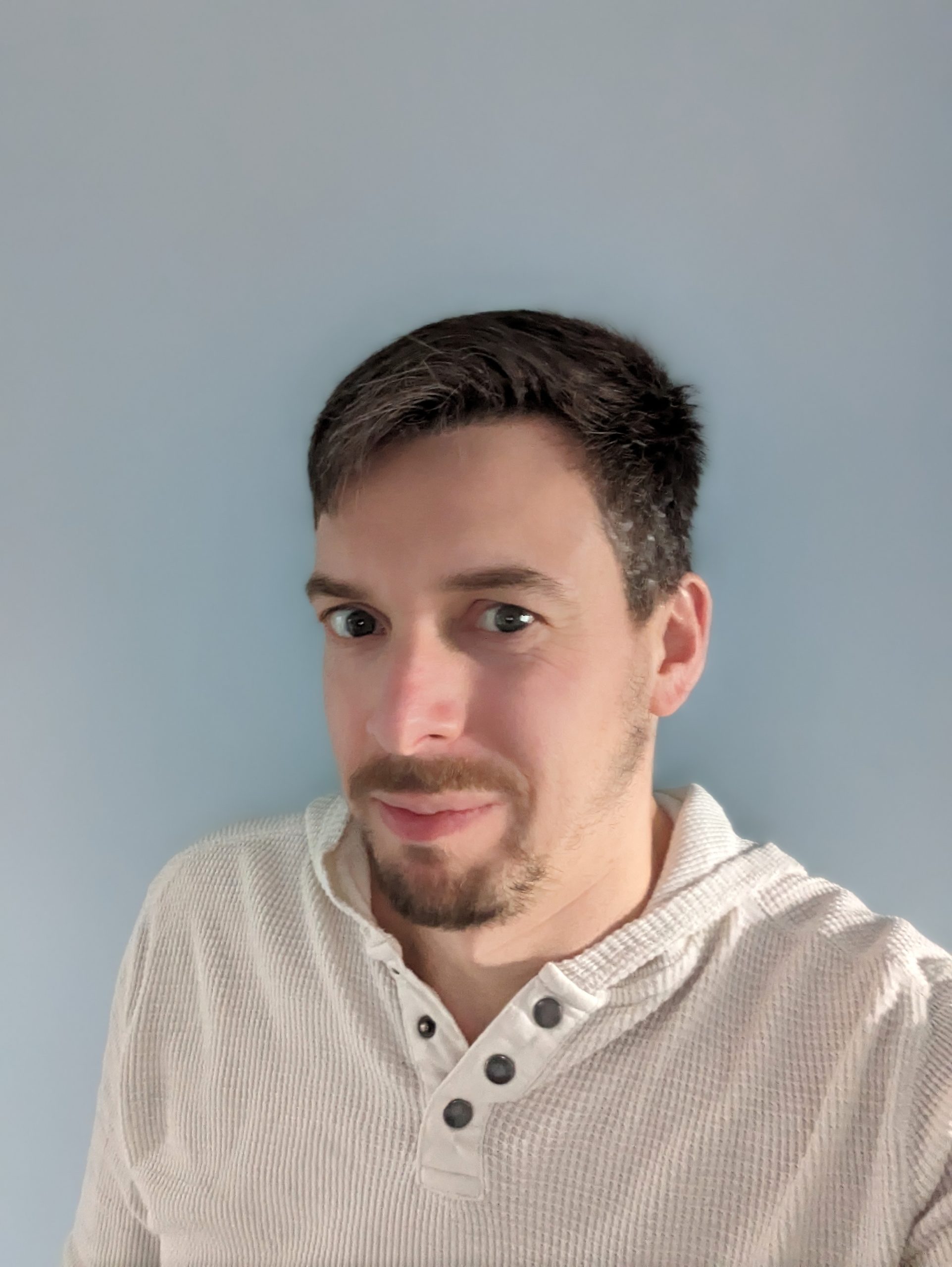 Photograph of white male with brown hair and beard wearing a cream top against a blue background