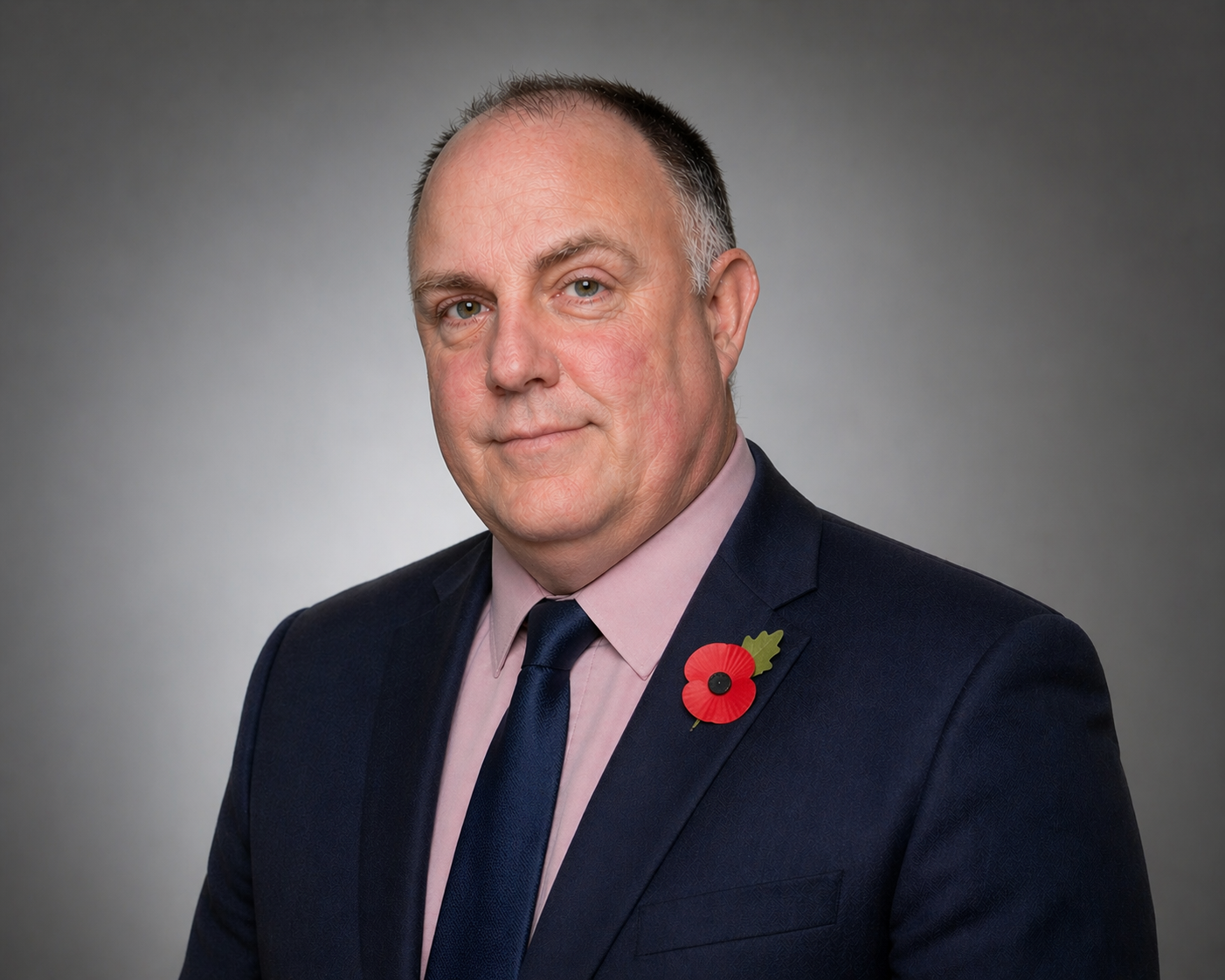 headshot of a white man in a black suit, pink shirt and tie with a poppy on his lapel