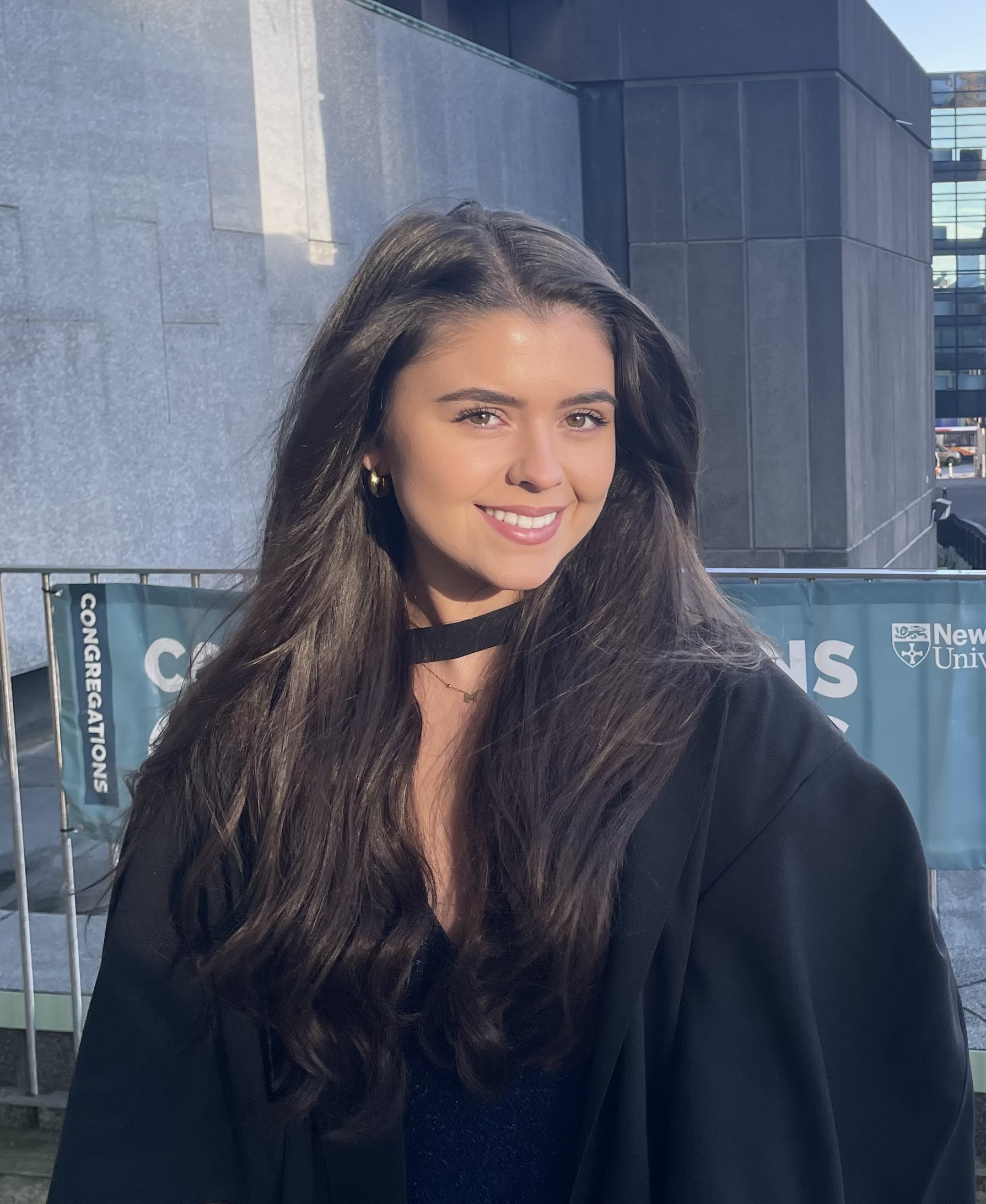 a white woman with long brown hair smiles whilst wearing a graduation gown